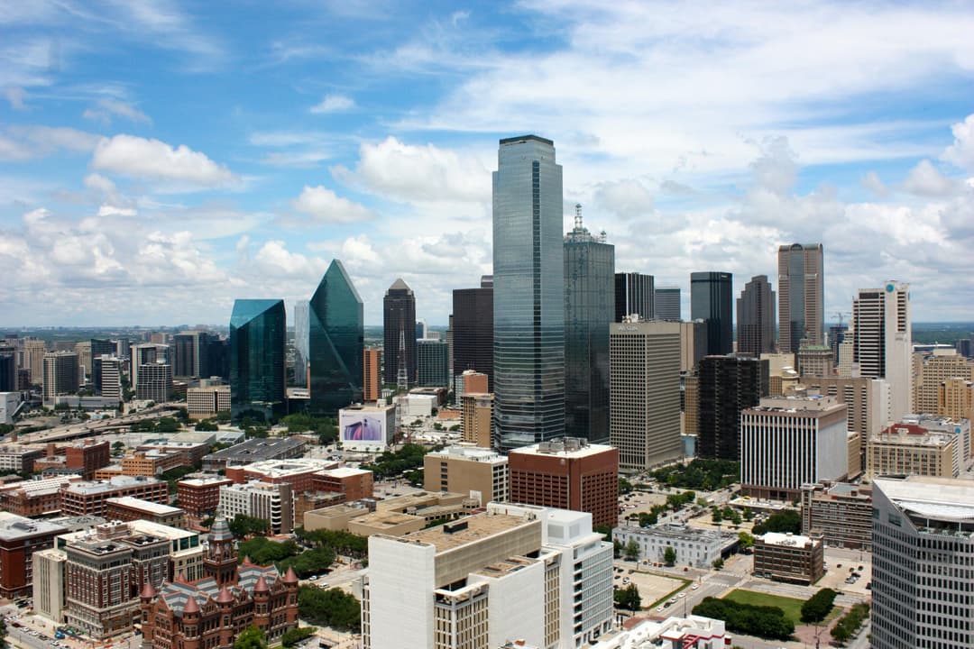 Aerial view of the downtown Dallas skyline with office buildings, city streets, and surrounding development under a blue sky.